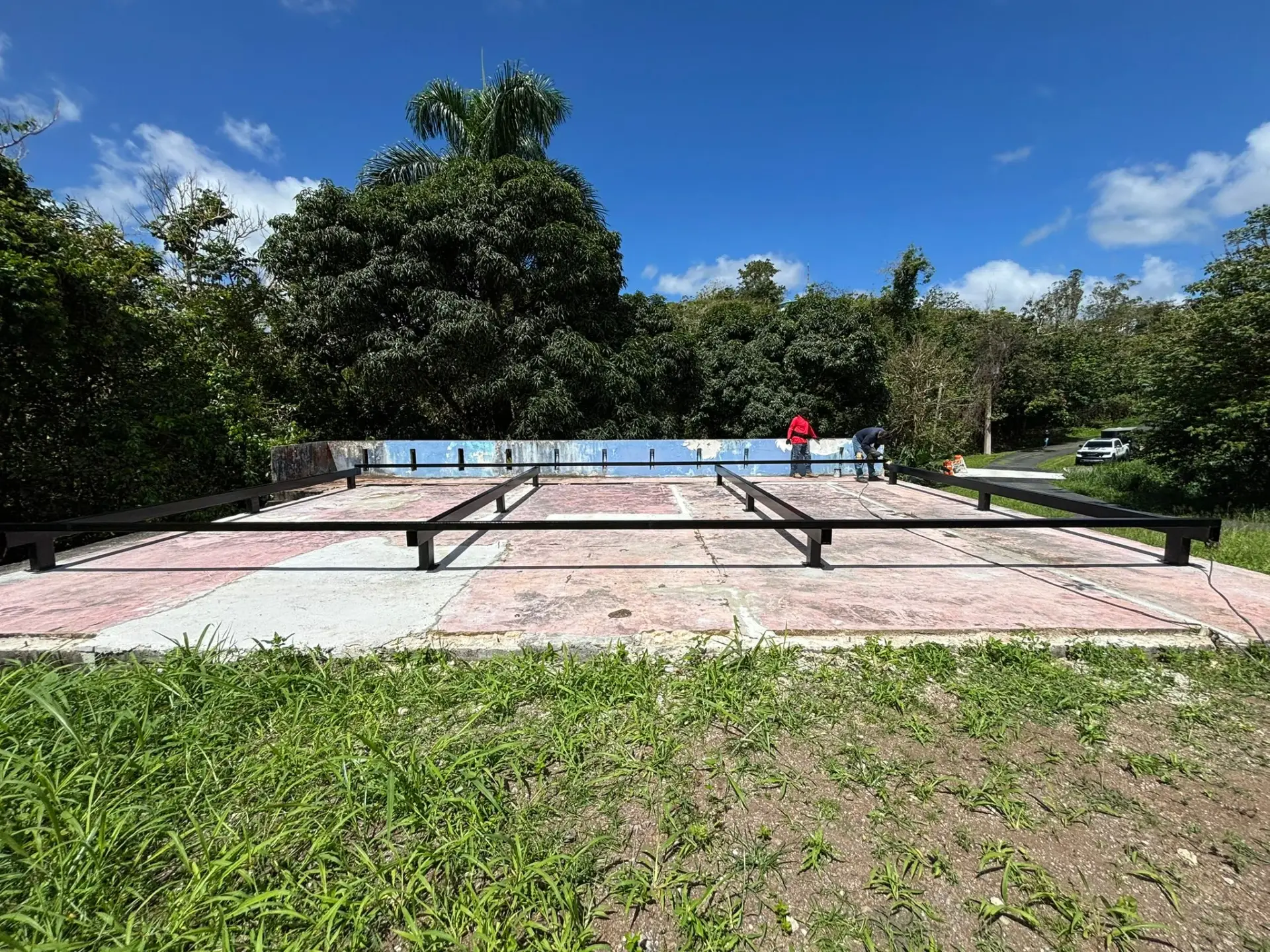 Steel foundation framing being installed on a prepared concrete pad surrounded by tropical greenery